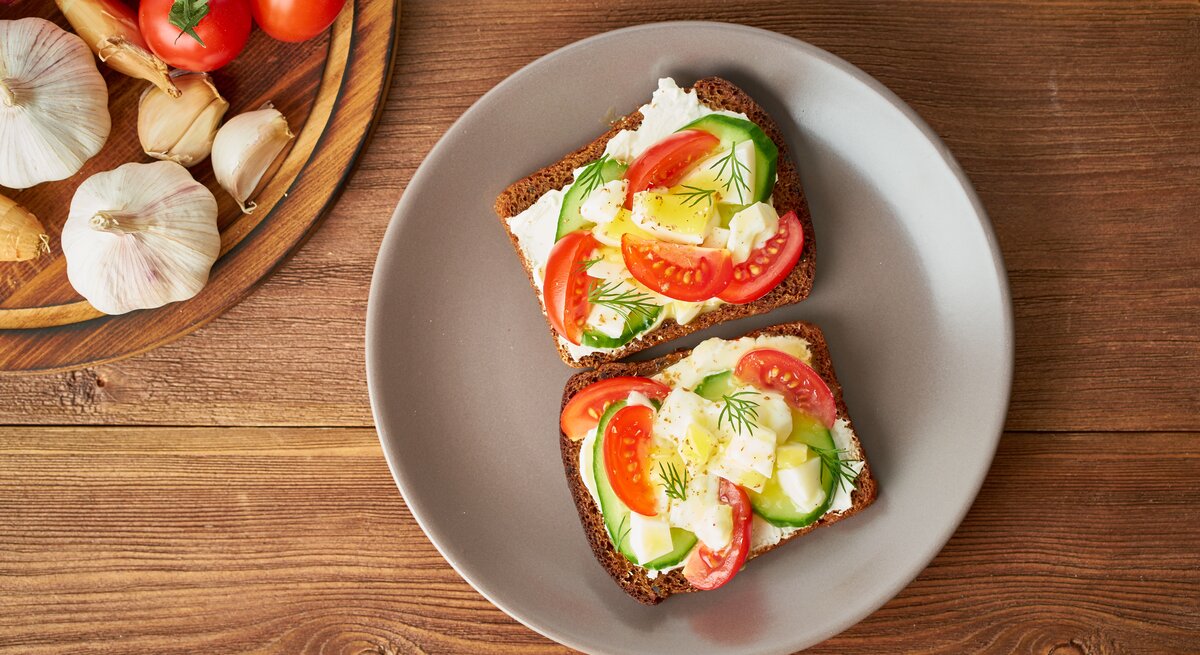 Draufsicht: Zwei Scheiben Brot mit Frischkäse, Gurken- und Tomatenscheiben und Ei liegen auf einem runden Teller. Der Teller steht auf einem Untergund aus Holz. Links daneben ist ein rundes Holzbrett teilweise zu sehen. Auf dem liegen Tomaten und Knoblauch.
