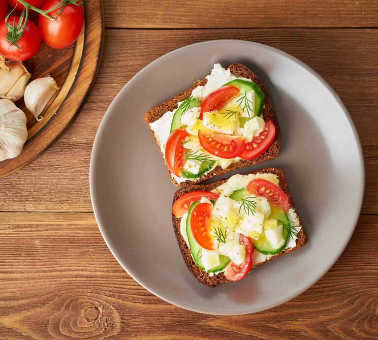 Draufsicht: Zwei Scheiben Brot mit Frischkäse, Gurken- und Tomatenscheiben und Ei liegen auf einem runden Teller. Der Teller steht auf einem Untergund aus Holz. Links daneben ist ein rundes Holzbrett teilweise zu sehen. Auf dem liegen Tomaten und Knoblauch. | © Shutterstock / Nata Bene
