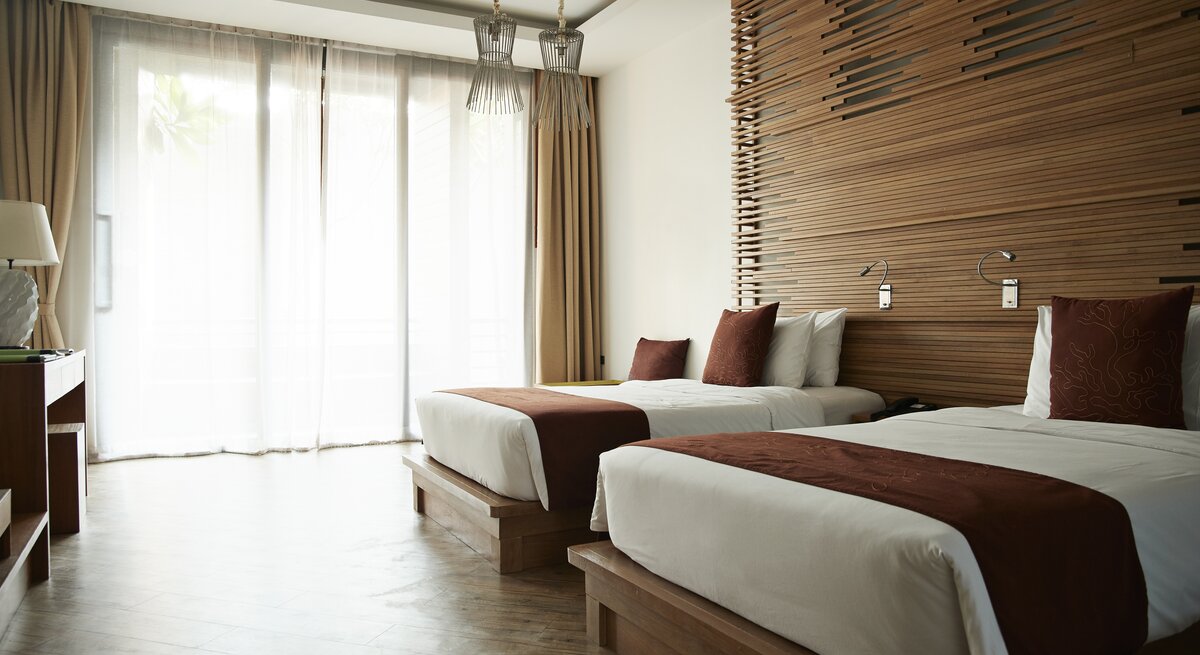 Modern gestaltetes Hotelzimmer mit zwei Einzelbetten. Jedes Bett ist mit einem weißen Laken und einem dunkelbraunen Bettschal am Fußende ausgestattet.