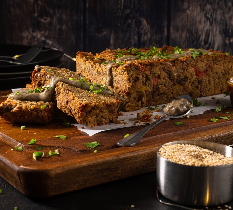 Auf einem großen Holzbrett liegt ein angeschnittener veganer Nussbraten mit etwas Soße beträufelt, drumherum eine Lichterkette und dunkler Hintergrund. | © Getty Images/ homydesign