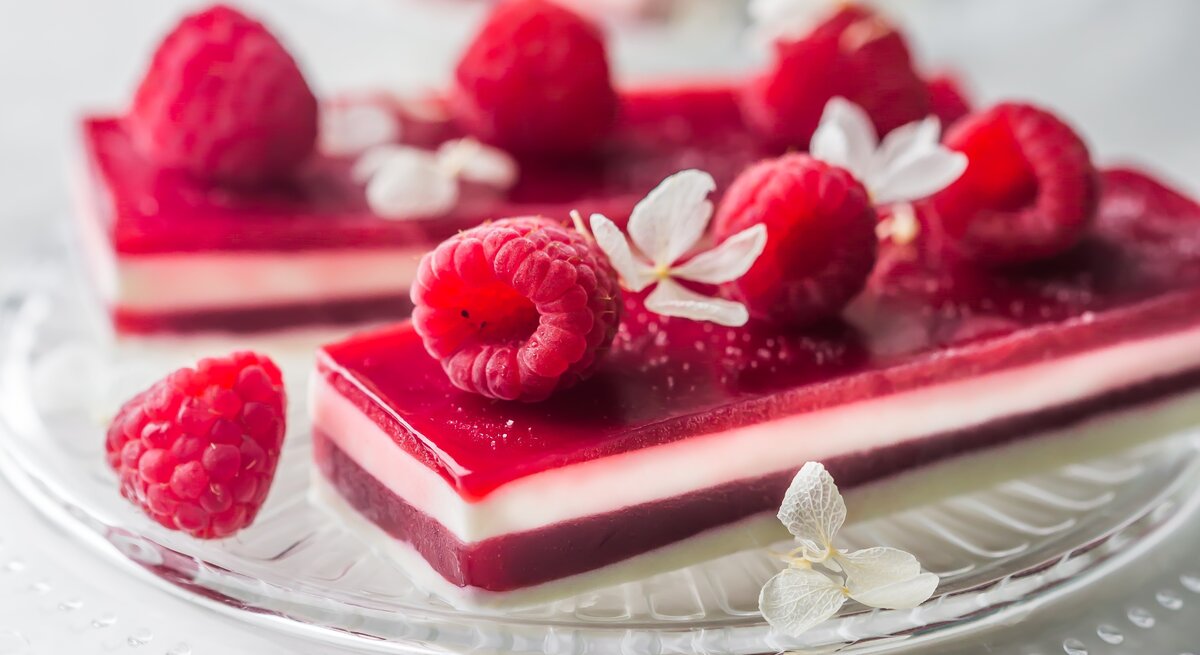 Panna-Cotta-Himbeer-Schnitten, garniert mit frischen Himbeeren und kandierten Früchten, serviert auf einem Kuchenteller auf hellem Untergrund. 