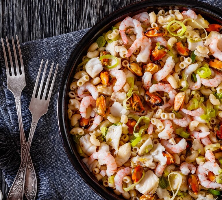 Eine schwarze Schüssel mit dem Salat auf einem dunklen Holztisch. Daneben noch einige Gewürze und eine graue Tischdecke mit zwei Gabeln. | © GettyImages/from_my_point_of_view