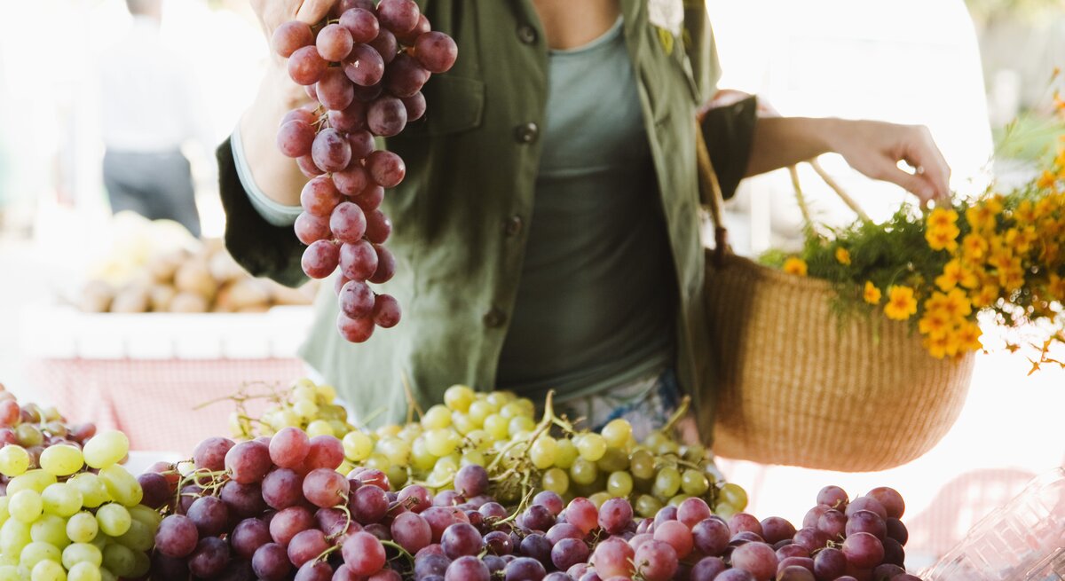 Das Bild zeigt eine Frau, die eine Traube roter Trauben über einem Marktstand hält, der mit verschiedenen Traubensorten gefüllt ist. Die Trauben erscheinen frisch und saftig, mit einem Farbspektrum von hellgrün bis dunkellila. Die Frau trägt ein grünes Oberteil und hat einen geflochtenen Korb über dem Arm, der mit Blumen geschmückt ist, was darauf hindeutet, dass sie frische Produkte und Blumen einkauft. Der Fokus liegt auf den Trauben und den Händen der Person, während der Hintergrund mit anderen Marktaktivitäten unscharf bleibt, was eine lebendige Marktatmosphäre vermittelt. 
