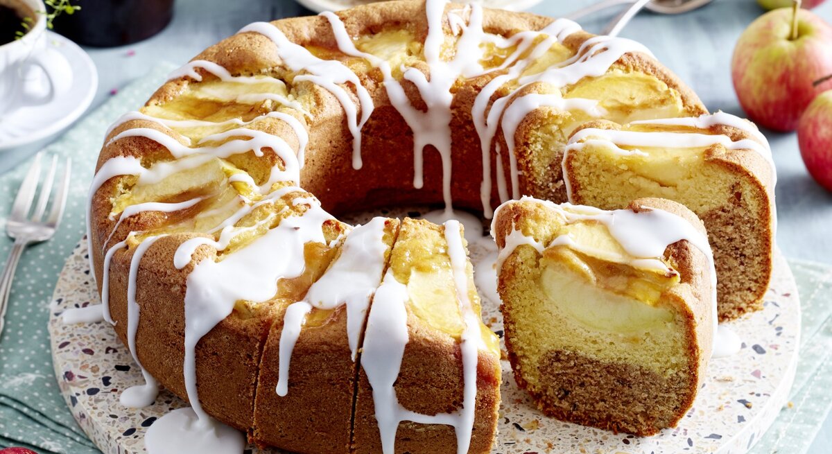 Angeschnittener Apfel-Kranzkuchen mit Zuckerguss auf Servierteller vor hellem Hintergrund mit Kaffeetassen und Äpfeln. 
