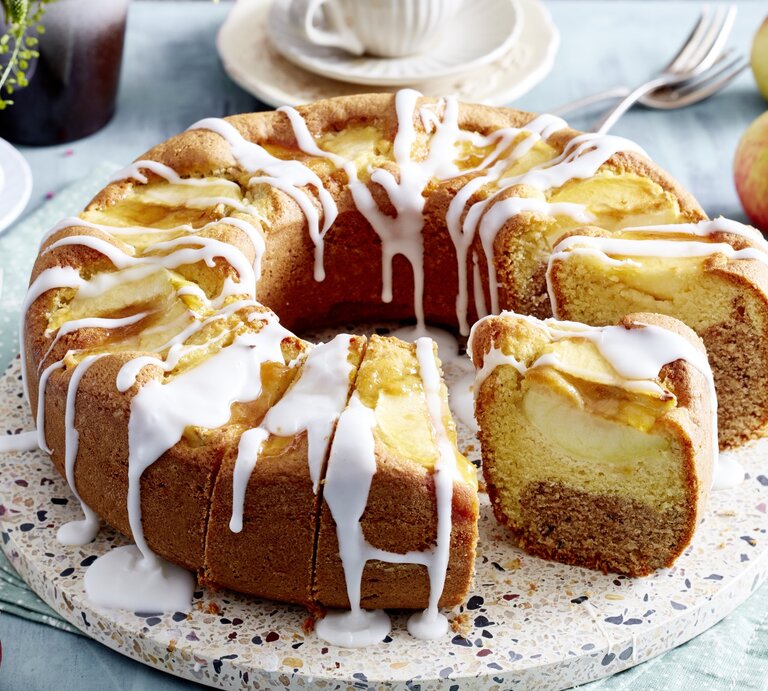 Angeschnittener Apfel-Kranzkuchen mit Zuckerguss auf Servierteller vor hellem Hintergrund mit Kaffeetassen und Äpfeln.  | © EAT CLUB / Isabell Triemer