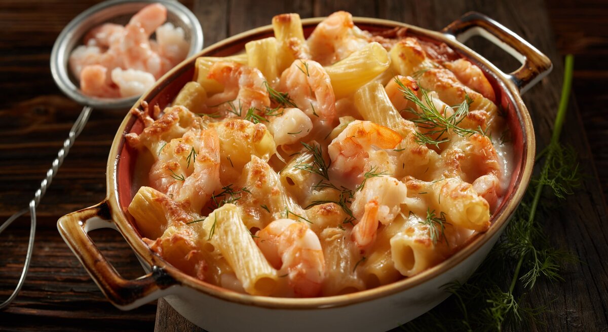 Frontalsicht: Auf einem dunklen Tisch steht eine dunkle Keramik-Auflaufform mit überbackener Pasta mit Dill-Schmand-Garnelen. Daneben liegt ein kleines Sieb mit rohen Garnelen.