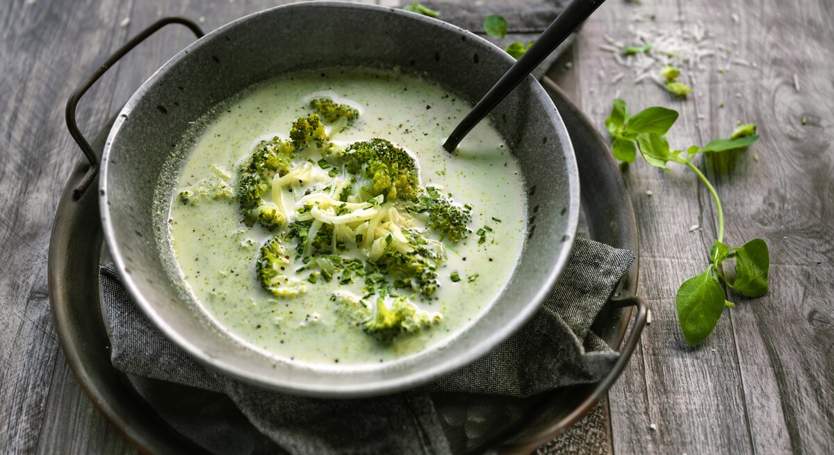 Auf einem rustikalen Holztisch sehe ich eine eiseren Pfanne in der eine schöne Schüssel aus Metall steht. In der Schüssel ist eine leuchtend grüne, cremig-pürierte Suppe, die mit ganzem Brokkoliröschen garniert ist. Die Suppe sieht sehr lecker aus! neben dem Ensemble liegt ein Zweig frisches Basilikum.