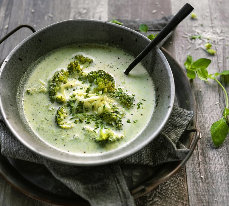 Auf einem rustikalen Holztisch sehe ich eine eiseren Pfanne in der eine schöne Schüssel aus Metall steht. In der Schüssel ist eine leuchtend grüne, cremig-pürierte Suppe, die mit ganzem Brokkoliröschen garniert ist. Die Suppe sieht sehr lecker aus! neben dem Ensemble liegt ein Zweig frisches Basilikum. | © lisegagne