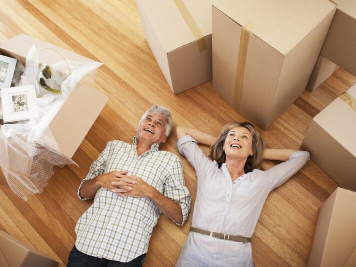 Reiferes Paar, das glücklich und erleichtert auf dem Boden ihres neuen Zuhauses liegt, umgeben von Umzugskartons und Einpackmaterial. | © iStock.com/Paul Bradbury
