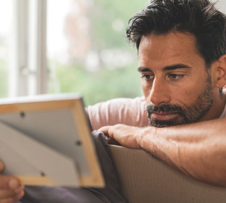 Nachdenklicher Mann mittleren Alters, der entspannt auf einem Sofa sitzt und aufmerksam ein Foto in einem Rahmen betrachtet. | © Getty Images / luza studios