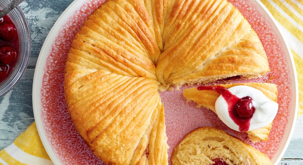 Auf einem Teller ist ein Wollknäuel-Butterkuchen angerichtet. Er ist mit Kirschen gefüllt und wird mit Sahne serviert.