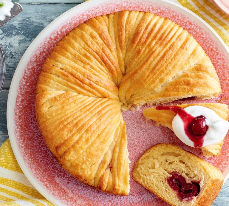 Auf einem Teller ist ein Wollknäuel-Butterkuchen angerichtet. Er ist mit Kirschen gefüllt und wird mit Sahne serviert. | © EAT CLUB / Isabell Triemer