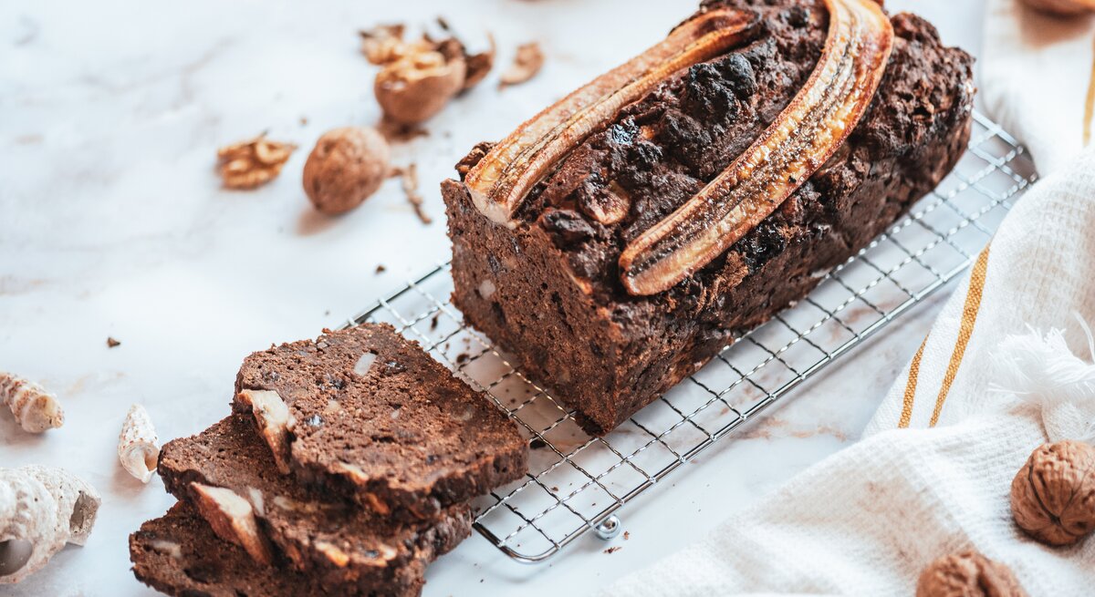 Ein angeschnittenes Schoko-Bananenbrot auf einem Kuchengitter, drumherum liegen geknackte Walnüsse.