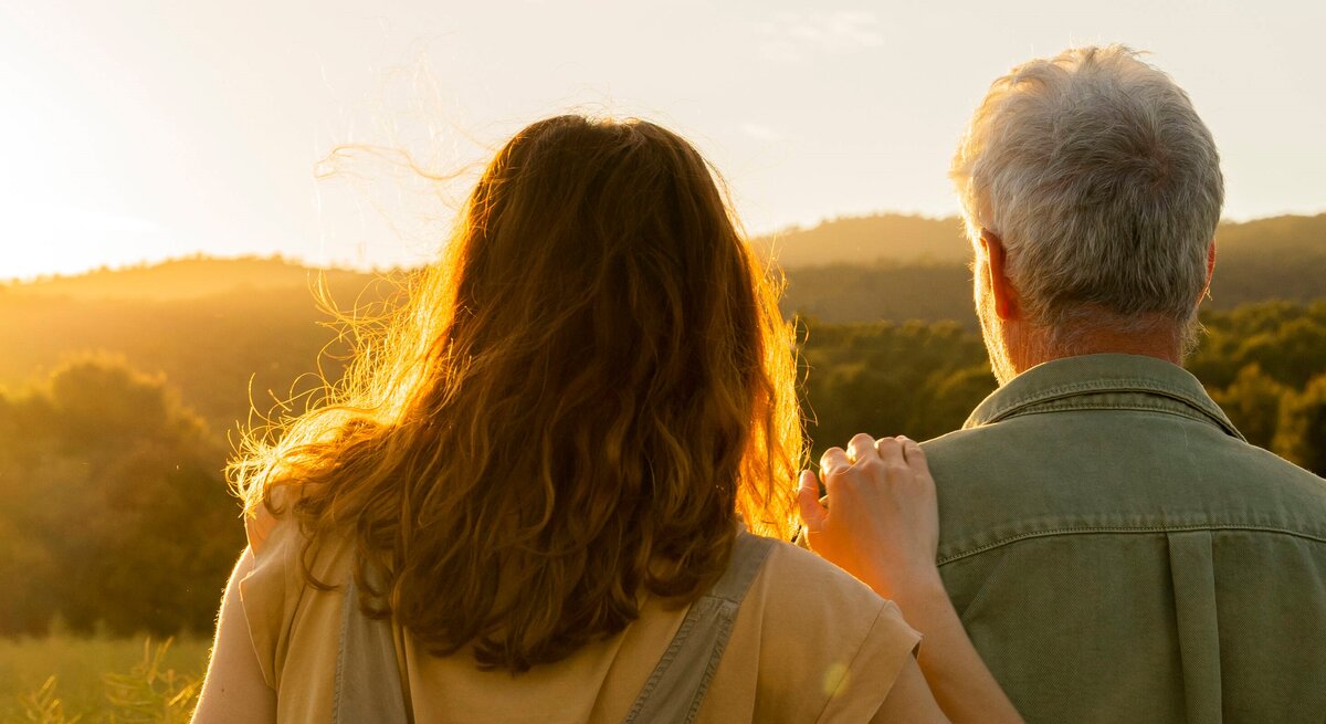 Zwei Personen von hinten, die gemeinsam in die Ferne blicken. Die Szene ist bei Sonnenuntergang oder Sonnenaufgang aufgenommen. Die Person auf der linken Seite hat schulterlanges, braunes Haar, während die Person auf der rechten Seite graues Haar hat. Die Person auf der linken Seite legt eine Hand auf die Schulter der anderen Person.