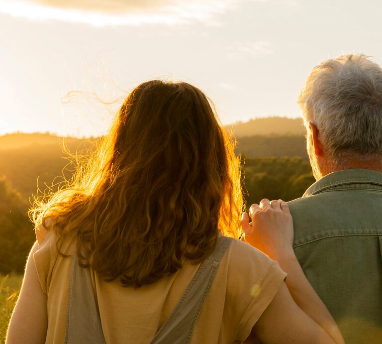 Zwei Personen von hinten, die gemeinsam in die Ferne blicken. Die Szene ist bei Sonnenuntergang oder Sonnenaufgang aufgenommen. Die Person auf der linken Seite hat schulterlanges, braunes Haar, während die Person auf der rechten Seite graues Haar hat. Die Person auf der linken Seite legt eine Hand auf die Schulter der anderen Person. | © IMAGO / Westend61