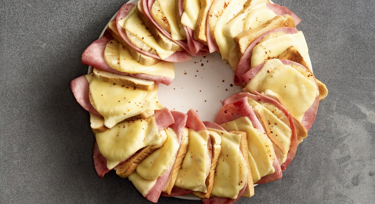 Hawaii-Toastbrot-Ring, bestehend aus abwechselnden Lagen von Gouda, Schinken, Ananas und Toastscheiben. In einem Ring aneinandergereit überbacken und mit Chiliflocken bestreut auf einem weißen Teller serviert. 