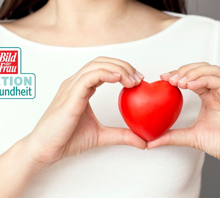 Eine Frau in weißem Oberteil hält vorsichtig ein rotes Herz in den Händen, das sie mit ihren Fingern sanft umschließt. Das Herzsymbol steht im Fokus des Bildes und symbolisiert die Bedeutung von Herzgesundheit. Das Logo "Bild der Frau AKTION Gesundheit" ist oben rechts im Bild platziert, was auf einen gesundheitlichen Kontext hinweist, möglicherweise im Rahmen einer Kampagne zur Förderung der Herzgesundheit. Die Frau ist nicht vollständig zu sehen, was den Fokus auf die Handlung der Herzpflege legt. | © ©istock/simarik [M]