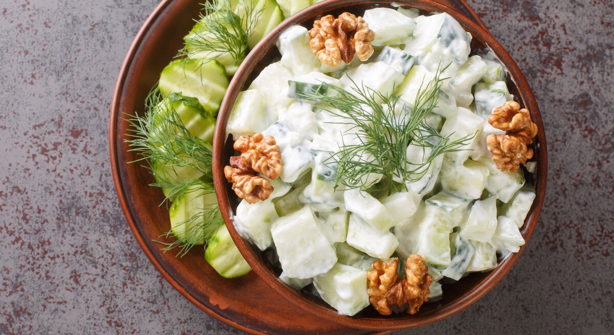 Bulgarischer Gurkensalat mit frischem Dill, Joghurt und ganzen Walnüssen, serviert in einer dunklen Keramikschale auf steinernem Untergrund. 