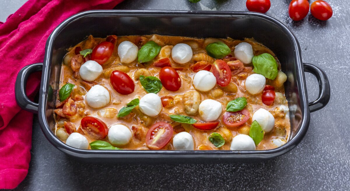 Schwarze Auflaufform gefüllt mit gebackenen Gnocchi mit Tomaten-Sahne-Soße und Mozzarella auf dunklem Untergrund. 