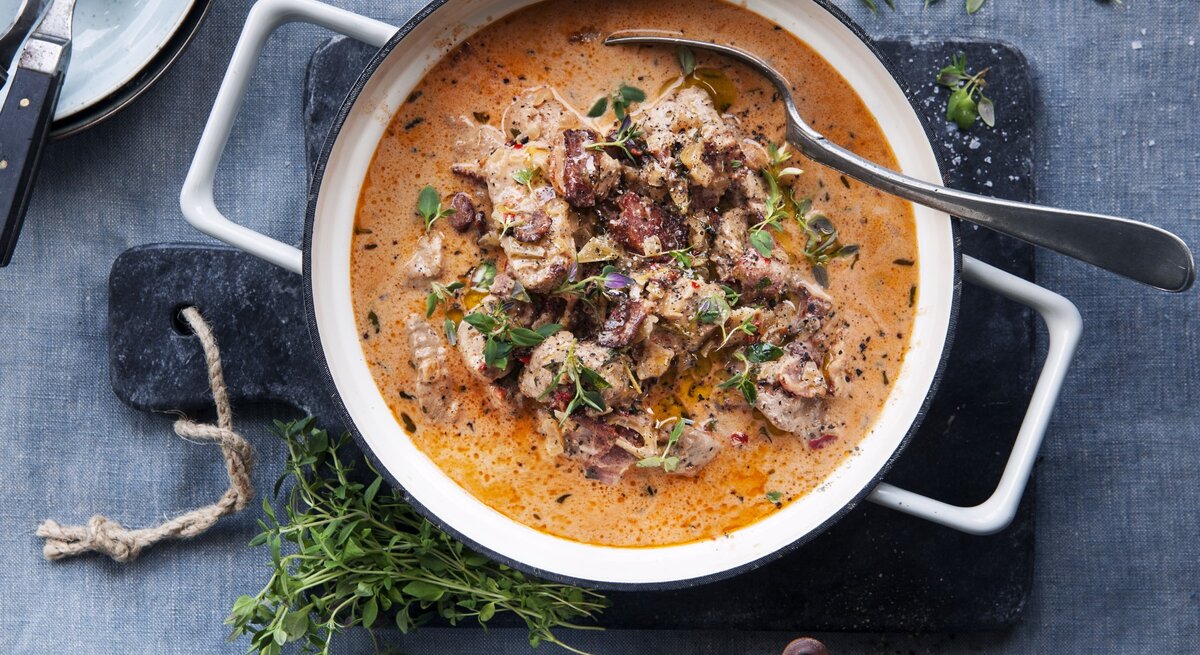 Eintopf mit Schweinefilet und getrockneten Tomaten, garniert mit frischem Thymian und angerichtet mit Sahne in einem großen Topf auf einer Schierferplatte. 
