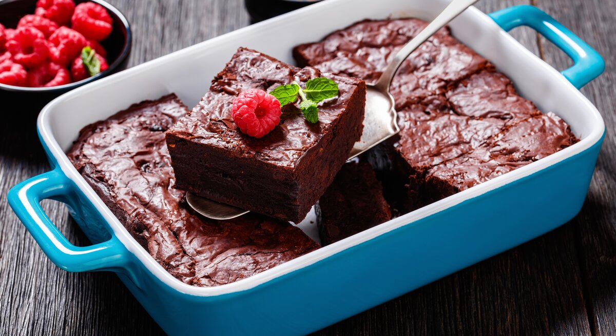 Himbeer-Brownies in blauer Auflaufform. Ein mit Himbeere und Minze belegtes Stück wird mit einem Tortenheber aus der Form gehoben. Daneben steht eine Schale mit Himbeeren. 