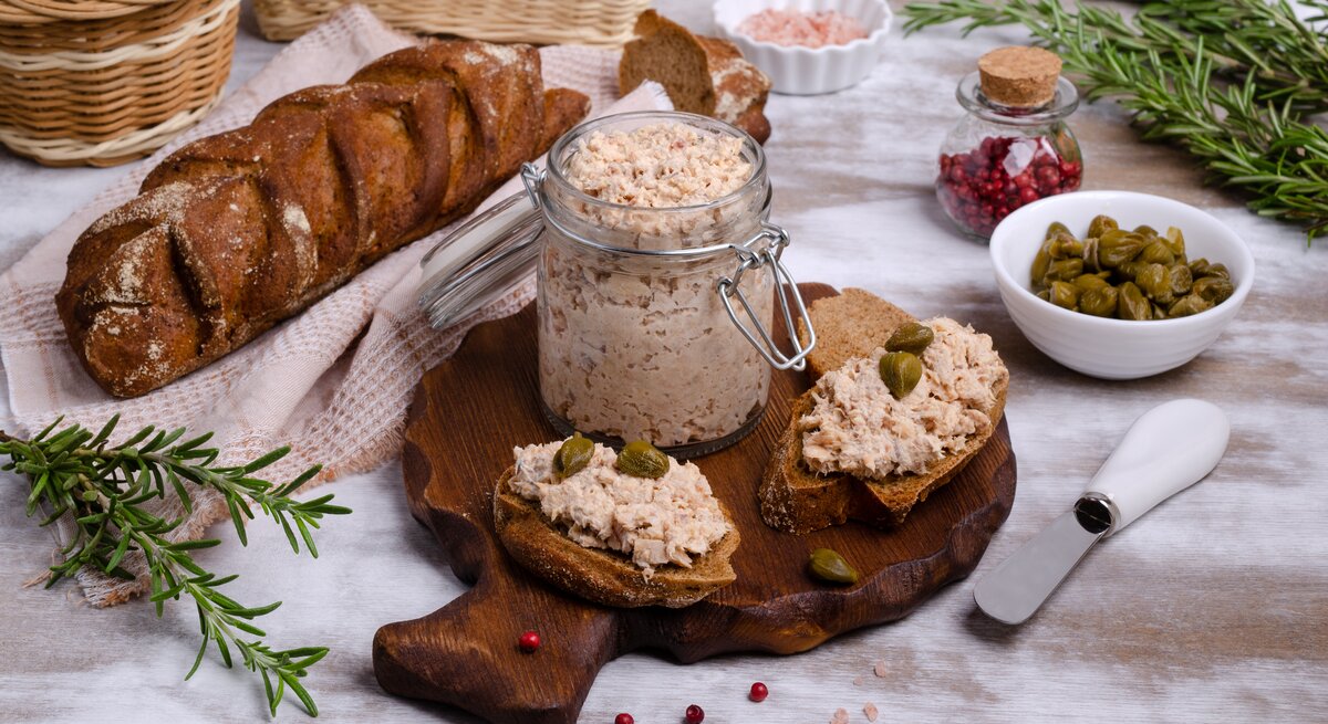 Frontal: Ein gedeckter Tisch mit Tischdecke, auf dem ein Glas Lachs-Rillette , eine Schale mit Kapern, ein Glas mit rosa Salz, ein Brotkorb, frischer Rosmarin und ein Brot liegen.