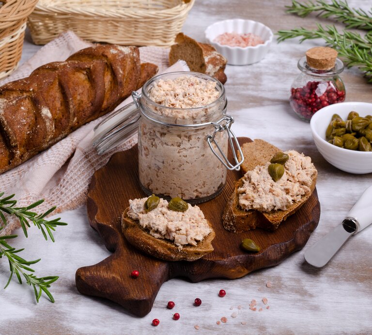 Frontal: Ein gedeckter Tisch mit Tischdecke, auf dem ein Glas Lachs-Rillette , eine Schale mit Kapern, ein Glas mit rosa Salz, ein Brotkorb, frischer Rosmarin und ein Brot liegen. | © Shutterstock / Veronika Idiyat