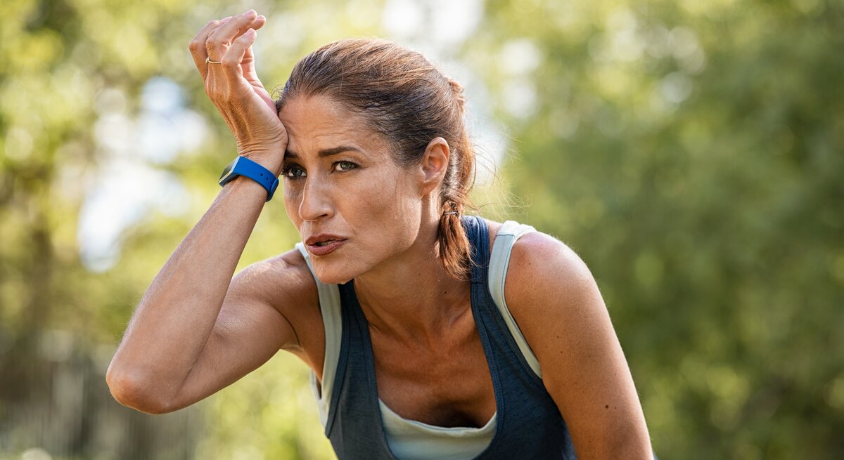 Eine erschöpfte sportliche Frau mittleren Alters mit einem Fitness-Tracker am Handgelenk wischt sich Schweiß von der Stirn, während sie sich in einer natürlichen grünen Umgebung erholt. Sie trägt ein ärmelloses Top und sieht aus, als hätte sie gerade ein intensives Training absolviert. Der Ausdruck auf ihrem Gesicht spiegelt Anstrengung und Konzentration wider.