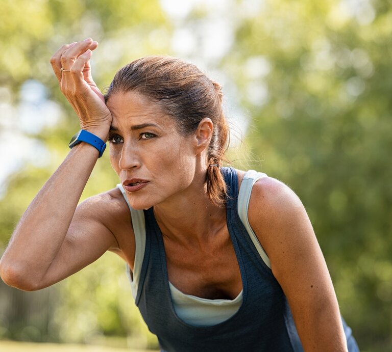 Eine erschöpfte sportliche Frau mittleren Alters mit einem Fitness-Tracker am Handgelenk wischt sich Schweiß von der Stirn, während sie sich in einer natürlichen grünen Umgebung erholt. Sie trägt ein ärmelloses Top und sieht aus, als hätte sie gerade ein intensives Training absolviert. Der Ausdruck auf ihrem Gesicht spiegelt Anstrengung und Konzentration wider. | © Getty Images/Ridofranz