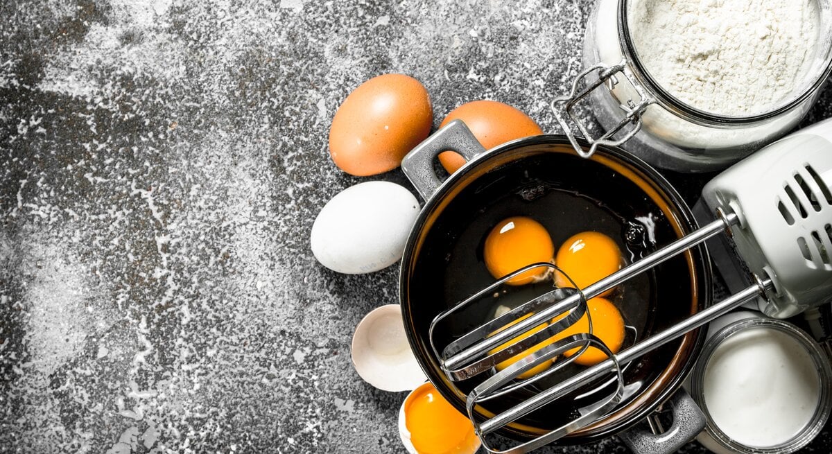 Draufblick: Auf einer dunklen bemehlten Arbeitsfläche steht eine Schüssel mit Eiern. Daneben sind ein Handmixer, Eier, Mehl, Sahne und Eierschalen zu sehen.