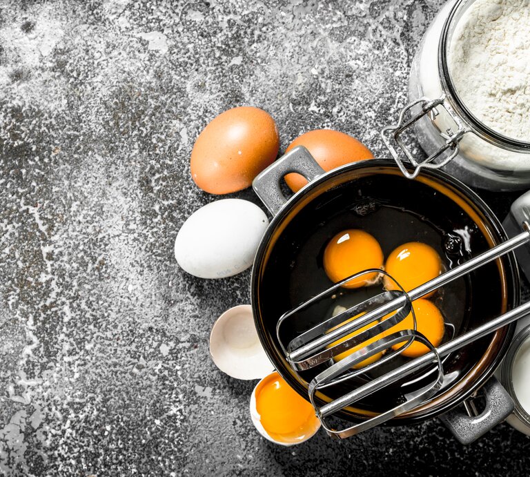 Draufblick: Auf einer dunklen bemehlten Arbeitsfläche steht eine Schüssel mit Eiern. Daneben sind ein Handmixer, Eier, Mehl, Sahne und Eierschalen zu sehen. | © Shutterstock / Chatham172