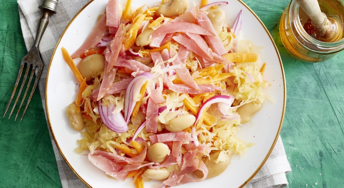 Auf einem Teller ist ein Sauerkraut-Salat mit weißen Bohnen angerichtet. Er ist mit Lachsschinken garniert.