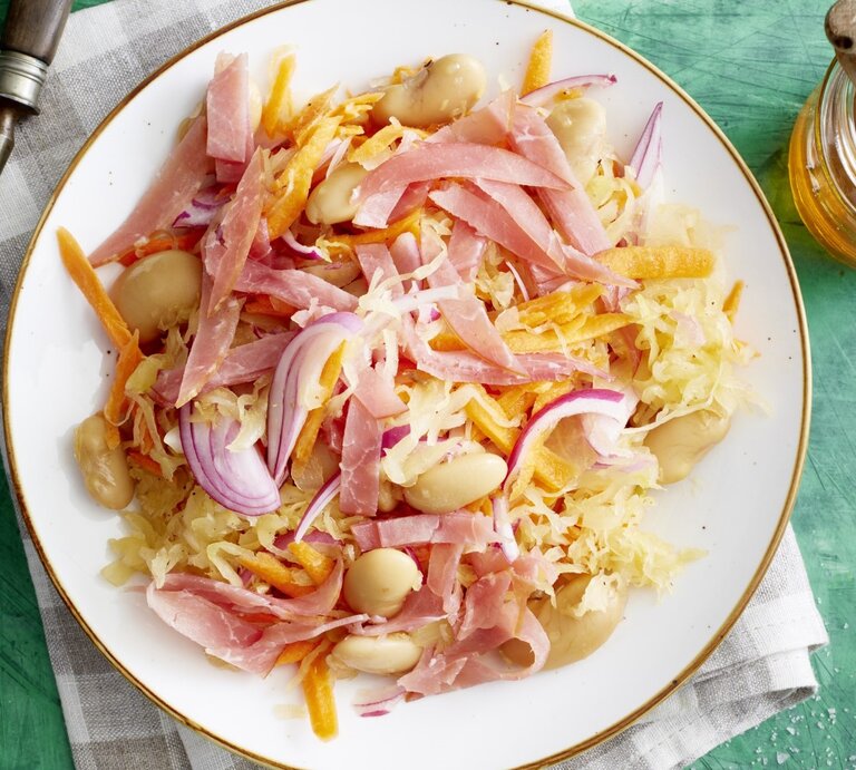 Auf einem Teller ist ein Sauerkraut-Salat mit weißen Bohnen angerichtet. Er ist mit Lachsschinken garniert. | © EAT CLUB / Isabell Triemer
