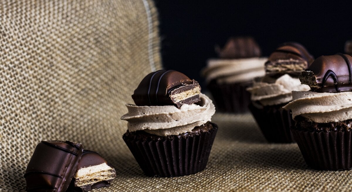 Kinder-Bueno Cupcakes mit Milchmädchencreme auf Jutebeutel vor schwarzem Hintergrund. Daneben Schokoriegel-Stücke. Frontalansicht. 