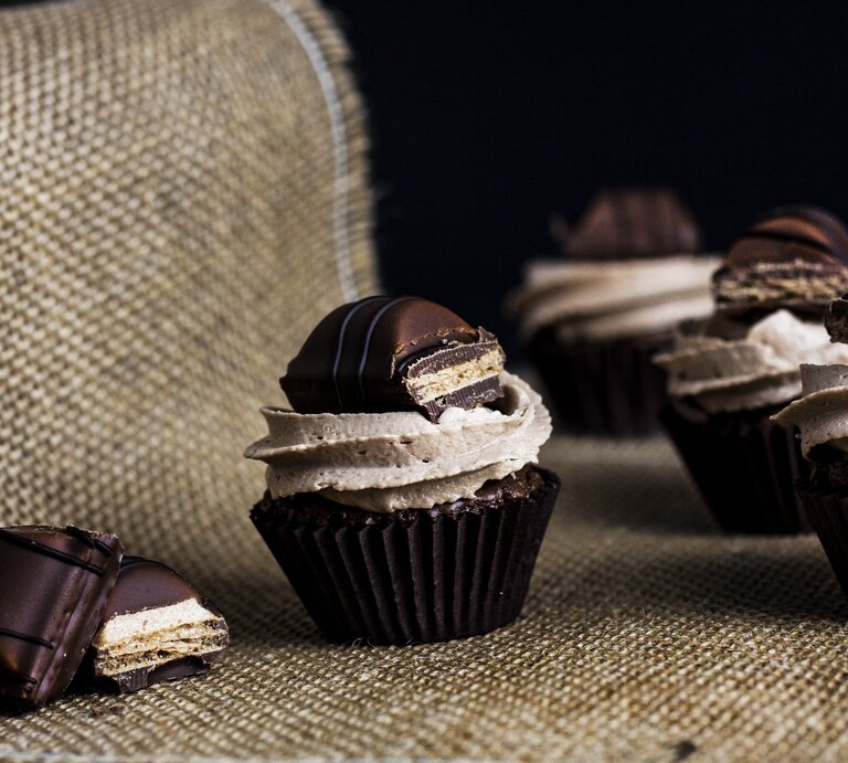 Kinder-Bueno Cupcakes mit Milchmädchencreme auf Jutebeutel vor schwarzem Hintergrund. Daneben Schokoriegel-Stücke. Frontalansicht.  | © shutterstock / Vitalie Stasii