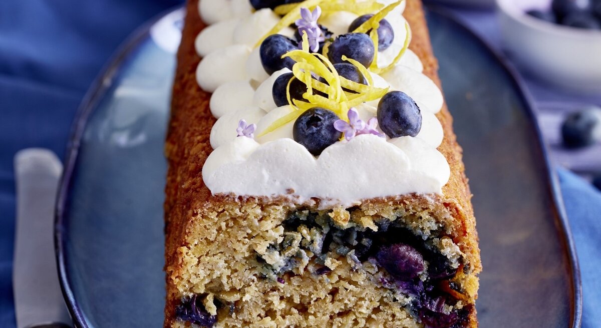 Auf einem Teller steht ein Heidelbeer-Haferflocken-Kuchen. Er ist mit Sahne und Blaubeeren dekoriert.