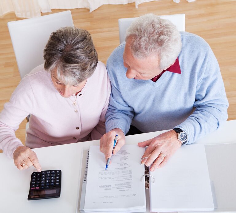 Ein älteres Paar sitzt an einem weißen Tisch, berechnet mit Taschenrechner und Unterlagen konzentriert seine Finanzen. | © iStock.com/AndreyPopov