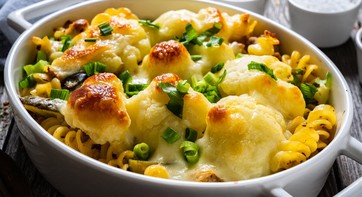 Schräg oben: ein überbackenes Blumenkohl-Gratin mit Hackfleisch und Käse in einer weißen Auflaufform steht auf einem Holztisch.