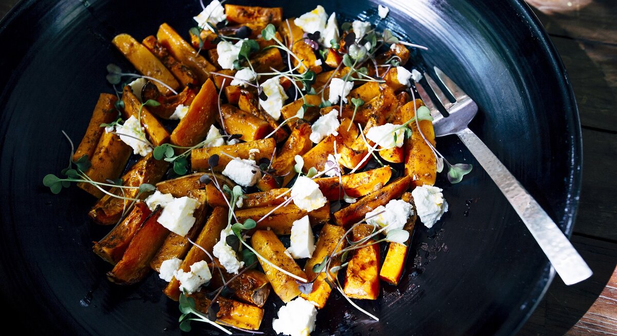 Eine schwarze Schüssel mit dem Süßkartoffelsalat und Feta nah fotografiert auf einem dunklen Holztisch.