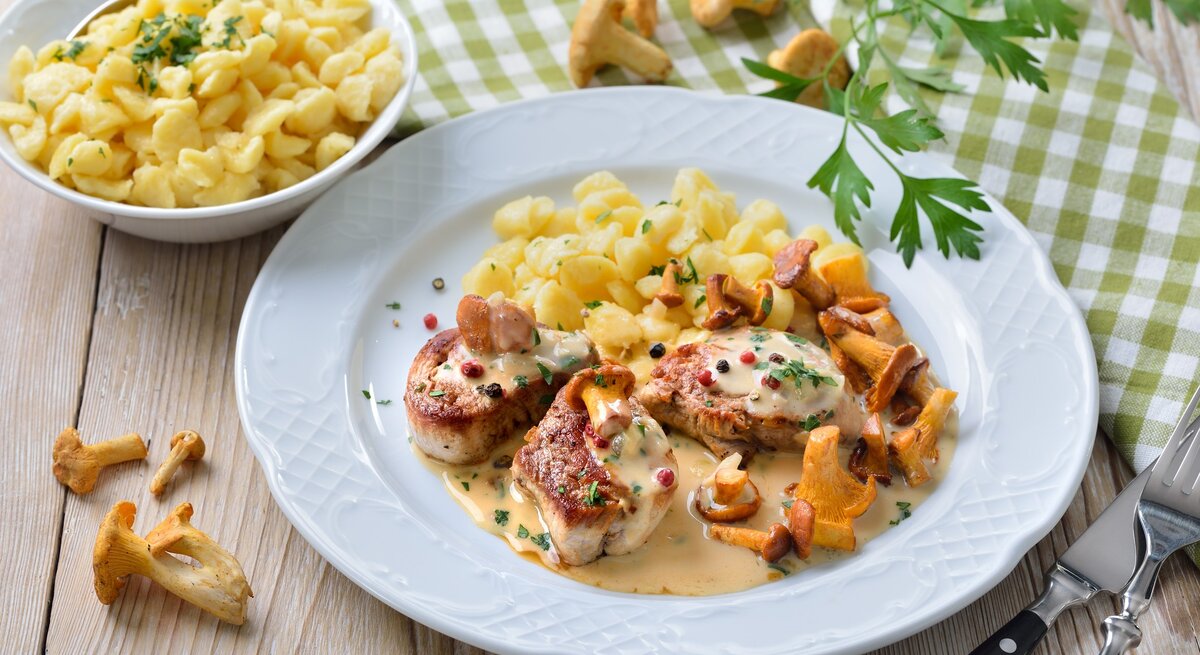 Schweinemedaillons mit Pfifferlingen in Rahmsoße mit buntem Pfeffer und Knöpfle auf weißem Teller und Holztisch. Daneben Schale mit Knöpfle. Petersilie und Pfifferlinge als Dekoration auf dem Tisch verteilt. Aufnahme aus der Vogelperspektive. 