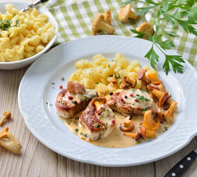 Schweinemedaillons mit Pfifferlingen in Rahmsoße mit buntem Pfeffer und Knöpfle auf weißem Teller und Holztisch. Daneben Schale mit Knöpfle. Petersilie und Pfifferlinge als Dekoration auf dem Tisch verteilt. Aufnahme aus der Vogelperspektive.  | © Getty Images / kabVisio
