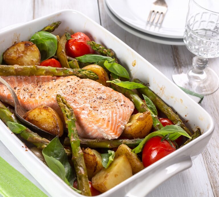 Eine weiße Auflaufform mit dem Ofenlachs auf einem weißen Holztisch. Daneben ein schickes Glas und zwei weiße Teller. | © Shutterstock/ Olga Nayashkova