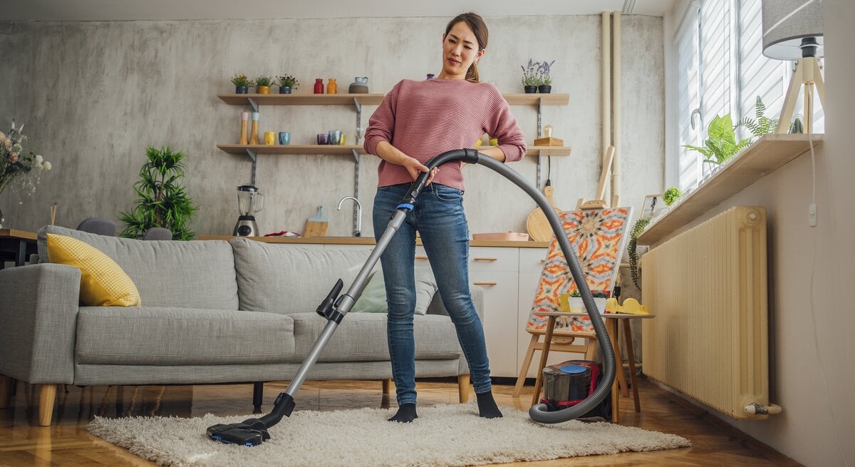 Eine Frau steht mit einem Staubsauger auf einem Teppich in einem gemütlichen Wohnzimmer und hält sich dabei den Rücken, als hätte sie Schmerzen. Sie trägt ein rosa Oberteil und Jeans und hat einen schmerzverzerrten Gesichtsausdruck, der auf Beschwerden beim Hausputz hinweisen könnte. Das Zimmer ist hell und einladend, mit einer Couch, Pflanzen, und einer modernen Einrichtung.