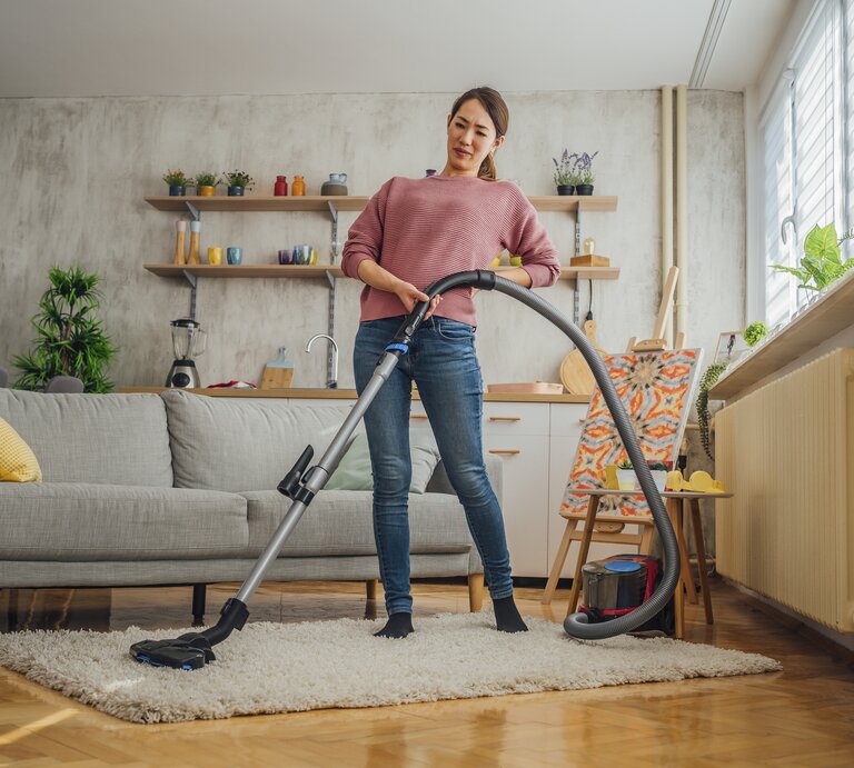 Eine Frau steht mit einem Staubsauger auf einem Teppich in einem gemütlichen Wohnzimmer und hält sich dabei den Rücken, als hätte sie Schmerzen. Sie trägt ein rosa Oberteil und Jeans und hat einen schmerzverzerrten Gesichtsausdruck, der auf Beschwerden beim Hausputz hinweisen könnte. Das Zimmer ist hell und einladend, mit einer Couch, Pflanzen, und einer modernen Einrichtung. | © Getty Images/LordHenriVoton