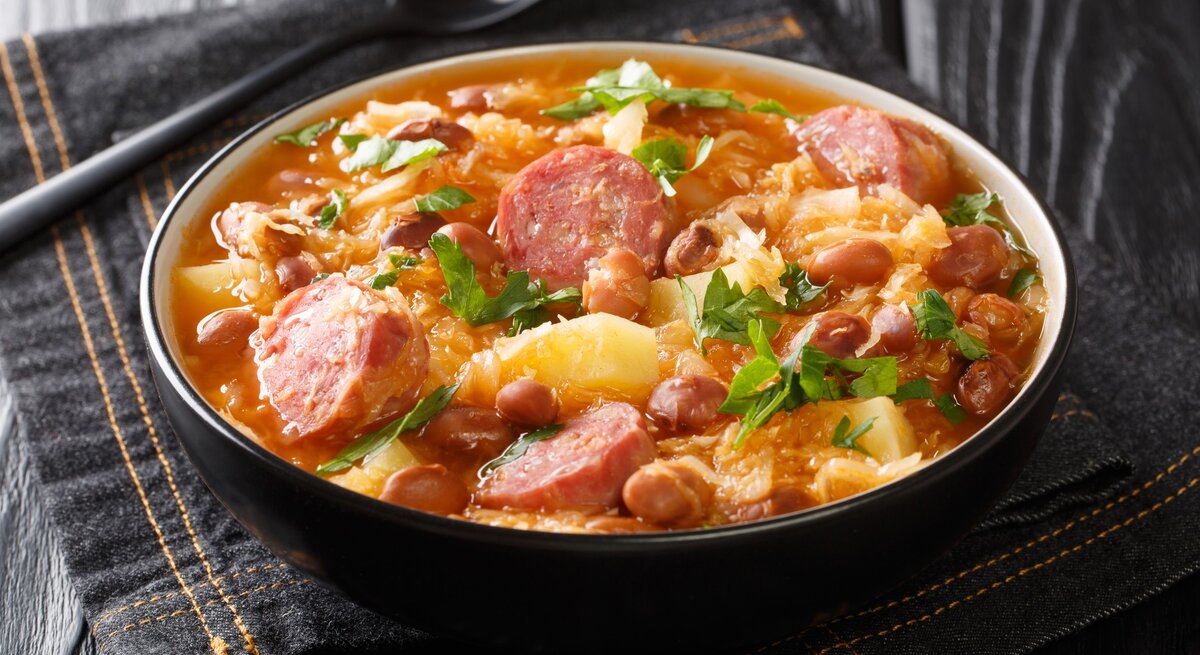 Sauerkraut-Eintopf mit Steckrüben und Räucherwurst in Suppenschüssel auf grauem Tuch. Daneben Suppenlöffel. Draufsicht.