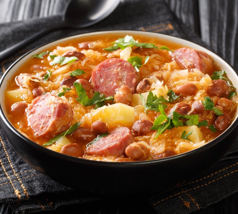 Sauerkraut-Eintopf mit Steckrüben und Räucherwurst in Suppenschüssel auf grauem Tuch. Daneben Suppenlöffel. Draufsicht. | © Getty Images / ALLEKO