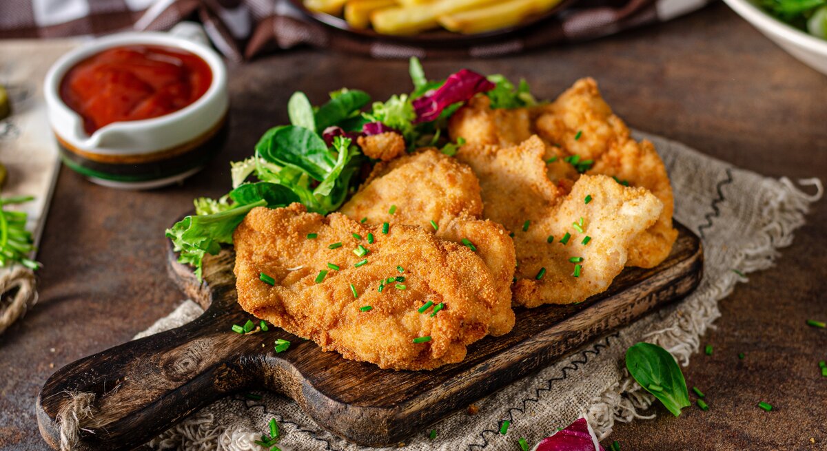 Ein Holzbrett mit drei Schnitzeln und etwas Schnittlauch auf einem dunklen Holztisch. Auf dem Tisch sind auch unscharf Pommes, Ketchup und Salat zu erkennen.