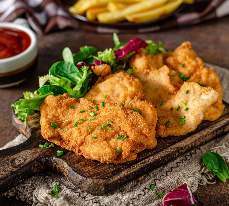 Ein Holzbrett mit drei Schnitzeln und etwas Schnittlauch auf einem dunklen Holztisch. Auf dem Tisch sind auch unscharf Pommes, Ketchup und Salat zu erkennen. | © Shutterstock/ Stepanek Photography