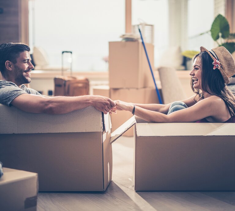 Junger Mann und junge Frau sitzen lachend jeweils in einem Umzugskarton in einer Wohnung | © iStock.com/skynesher