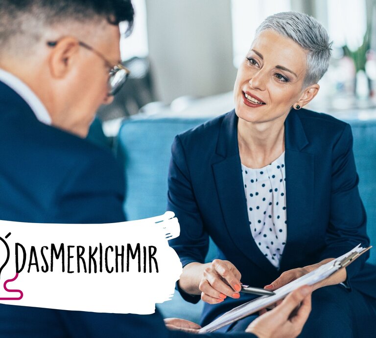 Das Bild zeigt ein professionelles Treffen zwischen einer Frau mit kurzen grauen Haaren und einem Mann, der von hinten zu sehen ist. Die Frau, die lächelnd und aufmerksam wirkt, trägt einen dunkelblauen Business-Anzug und ein gepunktetes Hemd. Sie hält einen Klemmbrett und scheint aktiv an dem Gespräch teilzunehmen oder dem Mann etwas zu erklären. Der Mann trägt einen dunklen Anzug und eine Brille. Im Vordergrund ist der Hashtag "#DAS MERK ICH MIR" neben einer Grafik einer Glühbirne zu sehen, | © iStock.com/filadendron